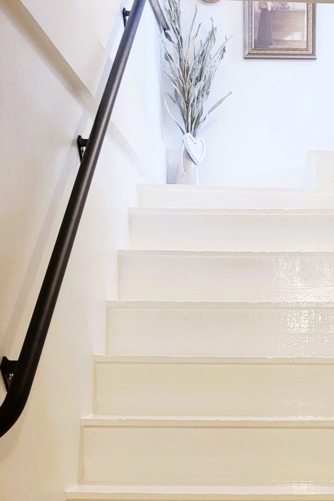 Bright white painted staircase with a custom matte-black handrail and a decorative vase at the top of the landing, showing the finished stairwell makeover.