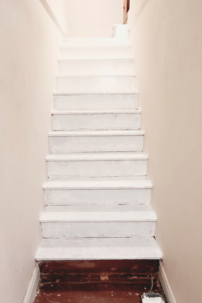 Stairwell after the first coat of white paint, showing partially painted wood treads with the bottom step still bare, creating a much brighter look during the makeover.