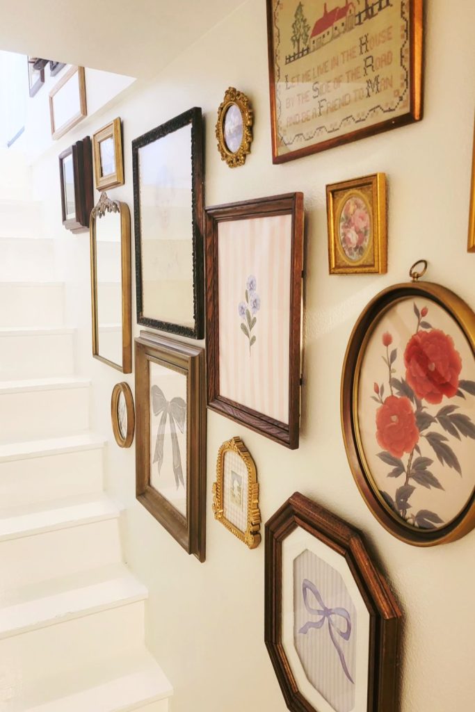 “White painted staircase with a vintage gallery wall of mixed frames, mirrors, and floral artwork arranged along the stairwell, showcasing the finished makeover.