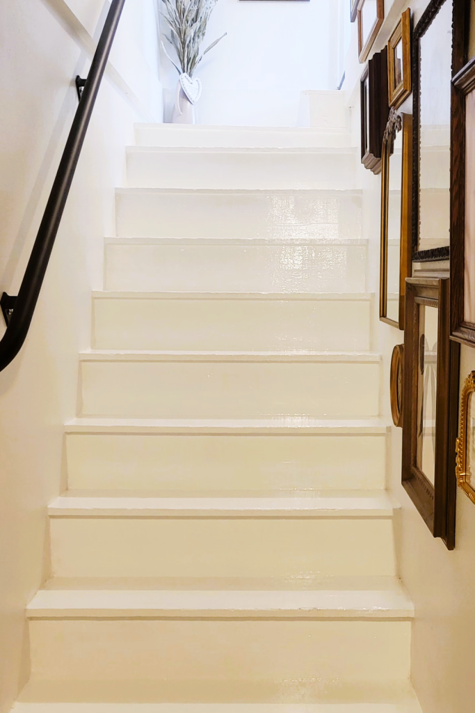Bright white painted staircase with a custom matte-black handrail and a vintage gallery wall of assorted frames lining the right side, showing the completed stairwell makeover.