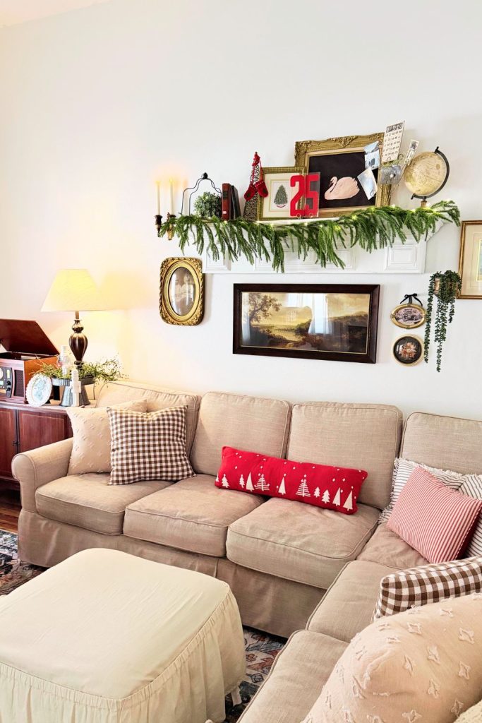 Cozy neutral living room with a sectional sofa, layered vintage artwork, and a Christmas garland shelf decorated with red and gold holiday accents.
