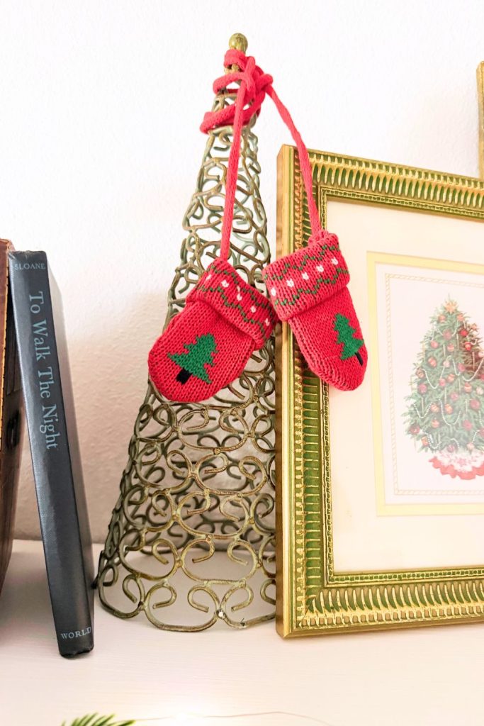 Small metal Christmas tree wrapped with red knit mittens beside a vintage gold frame and book, styled on a cozy holiday vignette shelf.
