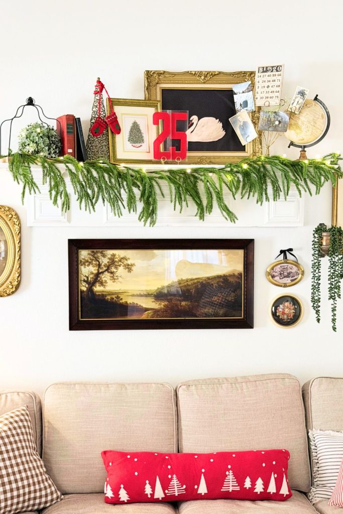 Layered farmhouse Christmas shelf styled with vintage frames, books, greenery garland, red “25” sign, and classic artwork above a beige sectional sofa.