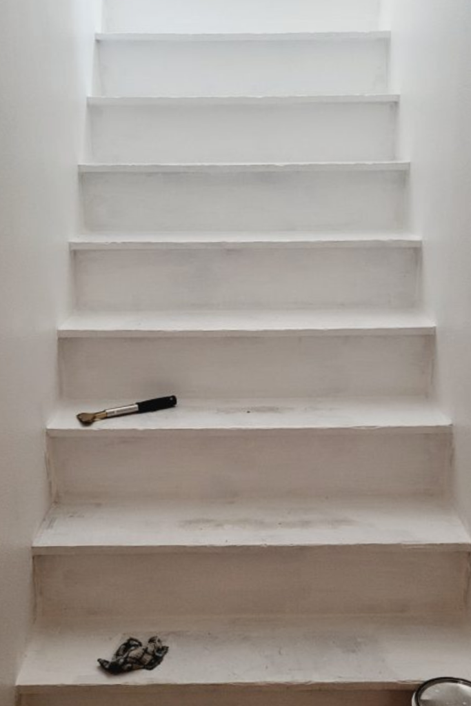 Stairwell after the first coat of white paint, showing partially painted wood treads with the bottom step still bare, creating a much brighter look during the makeover.