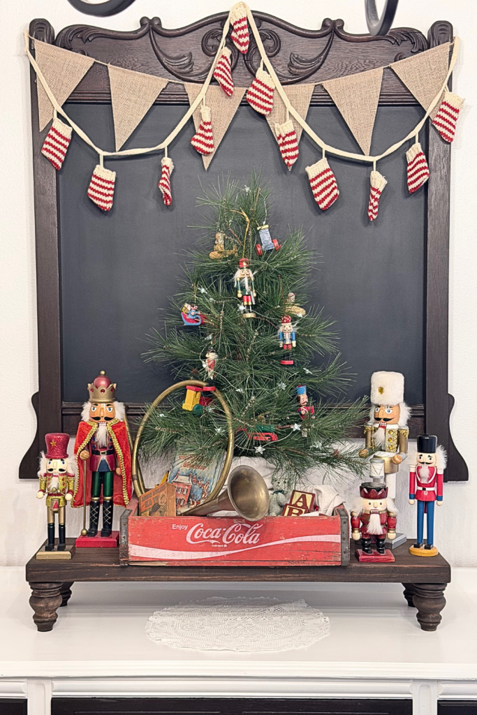 Styled Christmas buffet featuring a small pine tree decorated with nutcracker ornaments, vintage brass horn, red Coca-Cola crate, and striped mini-stocking garland hanging on a chalkboard backdrop.