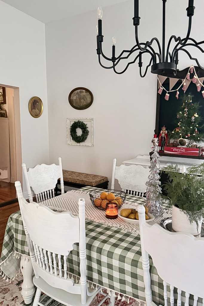 Vintage Christmas dining room with a green gingham tablecloth, white farmhouse spindle-back chairs, cozy holiday centerpiece with a lit candle, fruit bowl, and small evergreen plant. A silver tinsel tabletop Christmas tree and nutcracker vignette sit on a white buffet behind the table. Vintage wall art, a framed holiday wreath, and a black chandelier complete the cozy cottage-style Christmas decor.