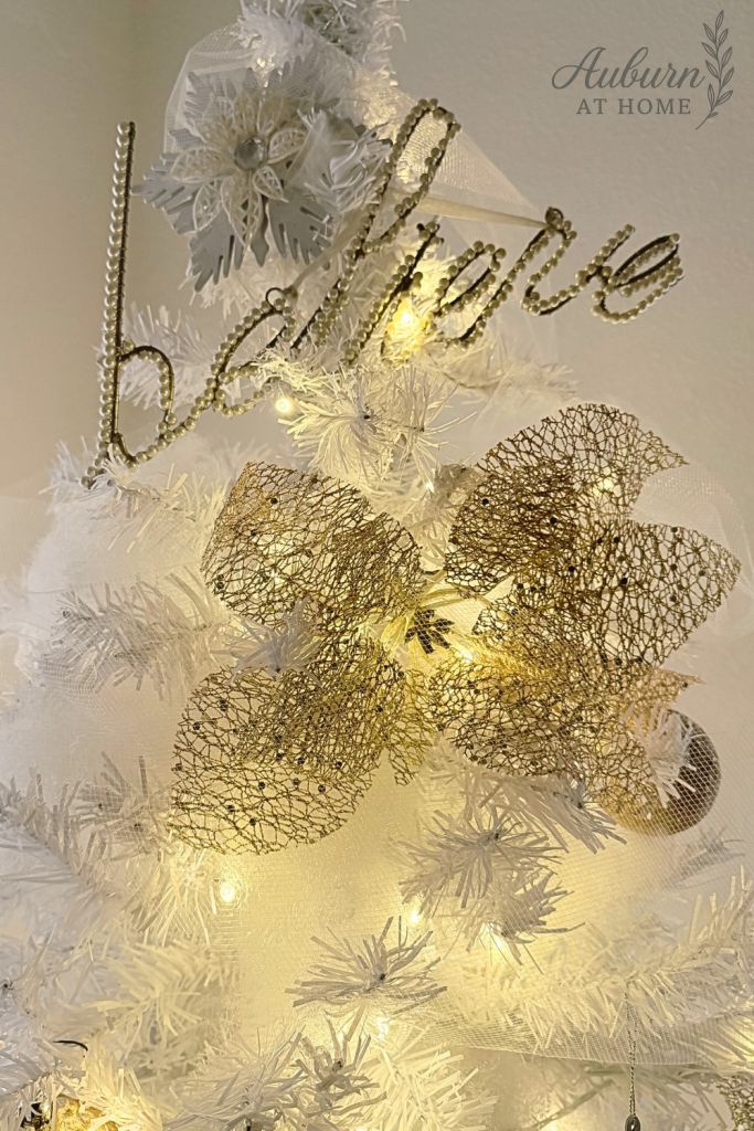 White Christmas tree decorated with gold mesh poinsettia and glitter ‘believe’ ornament glowing with warm lights.