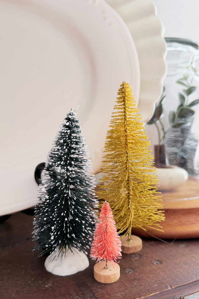 Small bottlebrush Christmas trees in black, gold, and pink arranged on a wooden surface in front of a white decorative plate for a simple holiday display.