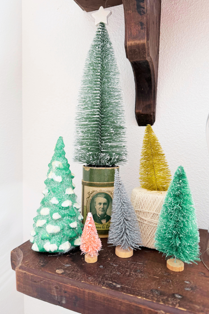 Colorful bottlebrush Christmas trees arranged on a wooden shelf, with one tree displayed on a vintage Edison tin and another elevated on a spool of twine.