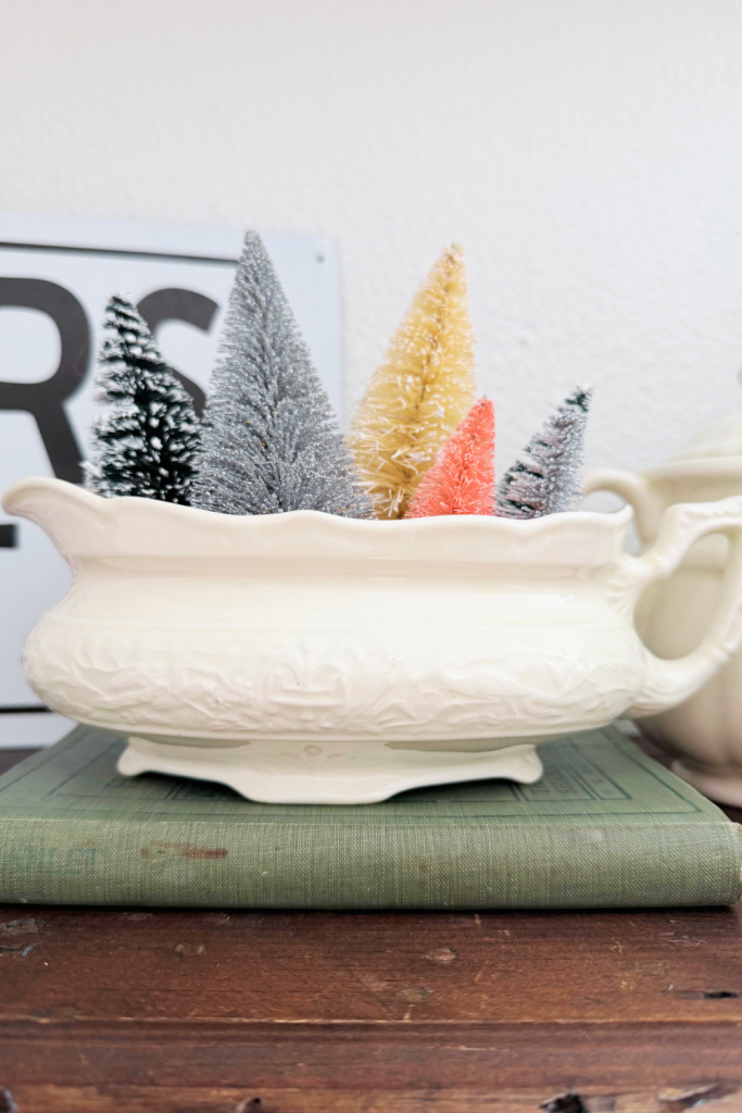 Colorful bottlebrush Christmas trees arranged inside a vintage white gravy boat, styled on top of a stacked book for a charming holiday display.