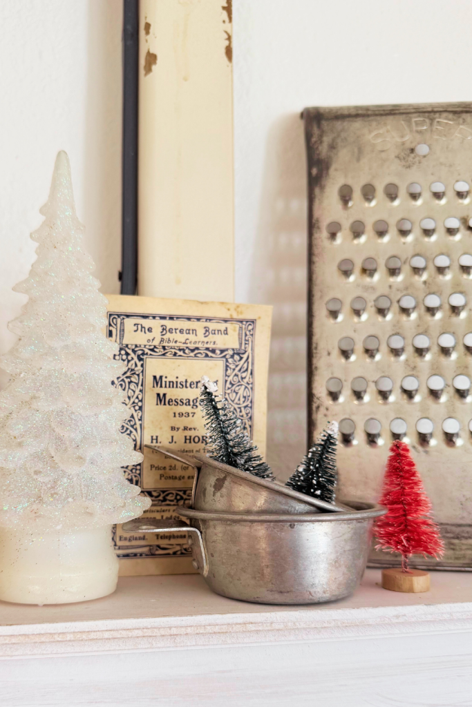 Vintage holiday vignette featuring small bottlebrush Christmas trees styled inside old metal cookware, with a 1937 booklet and antique grater in the background.