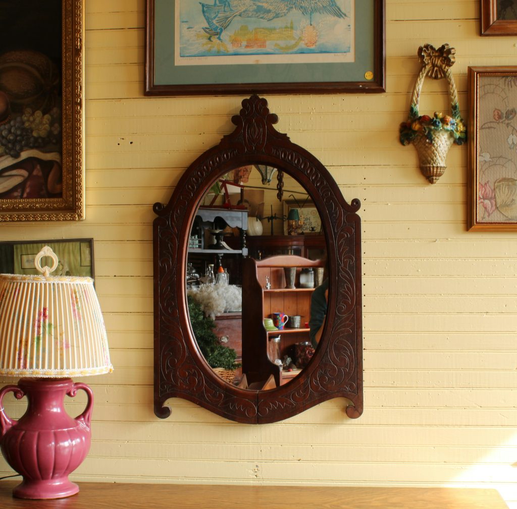 Antique carved wood wall mirror hung on a warm beadboard wall, surrounded by vintage artwork and soft cottage decor.